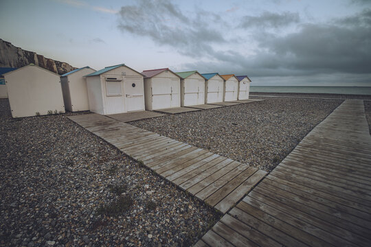 View of neat rows of beach huts with pastel-colored roofs stand on a shingle beach under a moody sky, connected by wooden walkways, Le Tr&Atilde;&copy;port, Normandy, France.