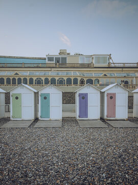 View of pastel beach huts stand in a neat row against a backdrop of grand architecture, pebbles scattered on the ground, sky above, Le Tr&Atilde;&copy;port, Normandy, France.
