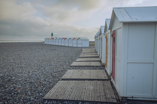 View of quaint, white beach huts stand in a neat row along a pebble beach under a cloudy sky, a wooden path leading to them, Le TrÃ©port, Normandy, France.