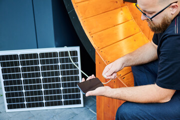 Cropped view of man connecting smartphone to photovoltaic solar panel. Integration of sustainable renewable energy into everyday life, demonstrating practical use of solar power for charging devices.