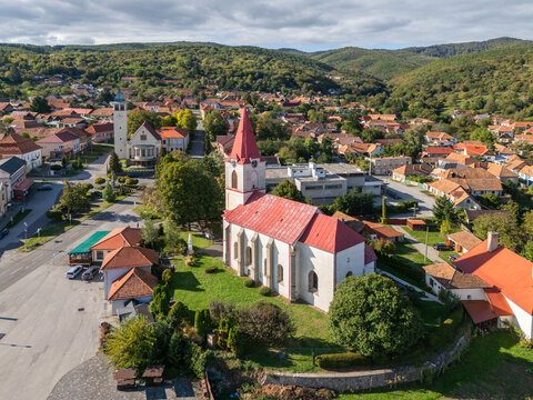Aerial view of a red-roofed church stands gracefully amidst a quaint village nestled in the embrace of rolling green hills, Torna&Auml;&frac34;a, Slovakia.