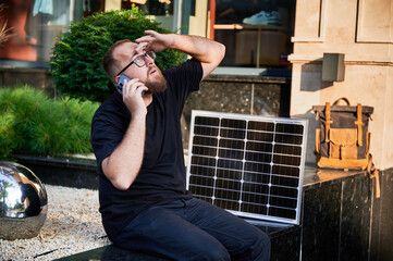 Bearded man talks on smartphone next to photovoltaic solar panel. Integration of sustainable renewable energy into everyday life, demonstrating practical use of solar power for charging devices.
