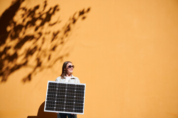 Woman holding solar panel for charging, stands against vibrant orange wall. Smiling girl wears sunglasses, white top. Artistic shadows cast by nearby trees branches on wall.
