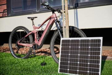 Pink electric mountain bike parked on grassy lawn, connected to solar panel for charging. Bike features rugged tires and positioned beside building, with its shadow cast on wall.