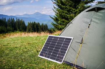 Green tourist tent pitched in grassy field with solar panel placed nearby. Solar panel highlights eco-friendly setup, surrounded by lush forest and rolling hills under blue sky.