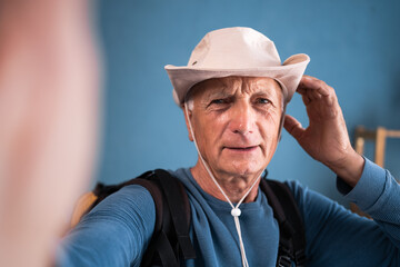 Elderly male traveler wearing hat and backpack taking selfie with smartphone at home before trip....