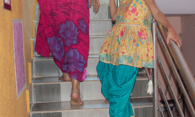 Two women, dressed in colorful traditional outfits with anklets and mehendi, gracefully ascend the...
