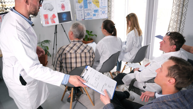 A group of people are sitting on chairs in the audience, and a doctor is standing in front of them. The doctor holds a tablet in his hands and hands it to one of the students