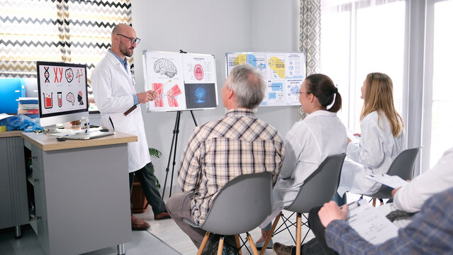 A group of researchers listens to a lecture by their colleague. A man in glasses and a white lab coat holds a presentation in front of a group of interns and residents