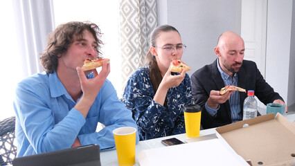 A group of happy people are eating pizza at an office table. Two men and a woman are having fun...