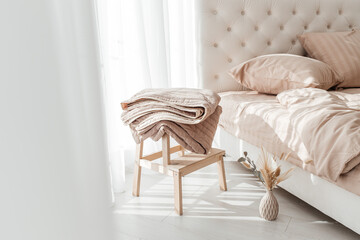 Cozy folded beige blanket on wooden stool next to a neatly made bed in morning sunlight