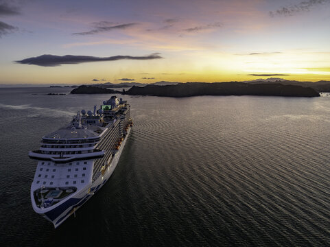 Mount Maunganui, New Zealand - 10 January 2024: Aerial view of a majestic cruise ship gliding through the serene waters towards distant islands under a sky painted with soft hues of pink and gold.