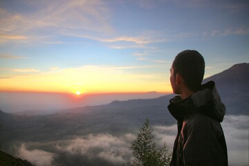 Silhouette of a man watching sunrise over mountains and clouds, ideal for travel, lifestyle, and...