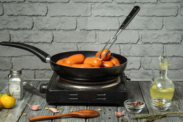 Juicy sausages are frying in a black pan on a stovetop, surrounded by garlic cloves, herbs, and a...