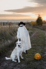 child in a white blanket, hat and black glasses walks his favorite white dog on Halloween. A young...