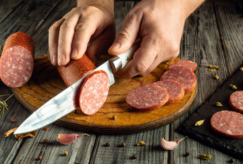 A person is skillfully cutting cured meat into thin slices on a wooden board. Garlic and spices add...