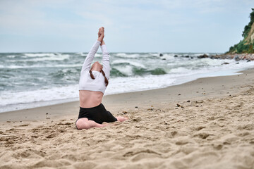 Young woman with brown hair performs yoga on baltic sea shore. Overcast sky and gentle waves create...