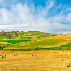 Obraz premium scenic agricultural landscape of summer season golden field with hay stacks during sunset or sunrise with green rural hills and beautiful mountains and clouds in evening sky on background