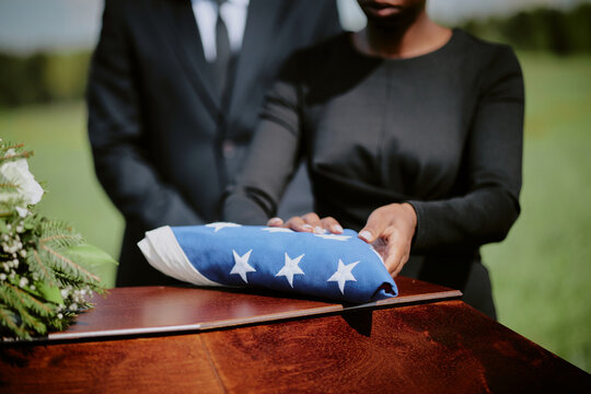 Black woman and Caucasian man standing at casket holding folded American flag during funeral ceremony outdoors, hands visible resting on flag, formal attire, solemn moment