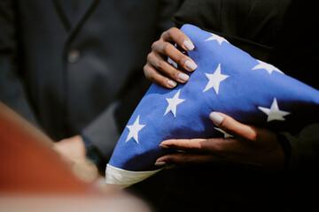 Black woman holding folded American flag during funeral ceremony, hands visible with manicured nails, standing beside Caucasian man in formal attire, honoring military service
