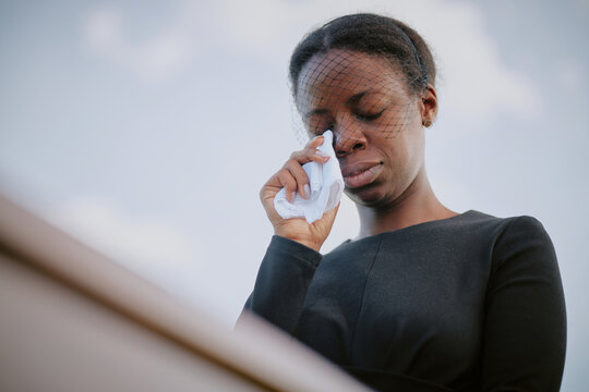 Black young adult woman wearing mourning attire standing outdoors wiping tears with tissue during funeral ceremony, eyes closed, expressing grief and sorrowful emotion