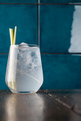 Refreshing cocktail with lemongrass stalks and ice cubes in a clear glass, served on a wooden table against a teal background, condensation on the glass, close-up