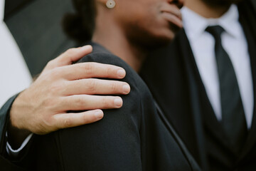Caucasian man comforting Black woman by placing hand on her shoulder during funeral, both wearing formal attire, partial faces visible, showing support and empathy in moment of grief