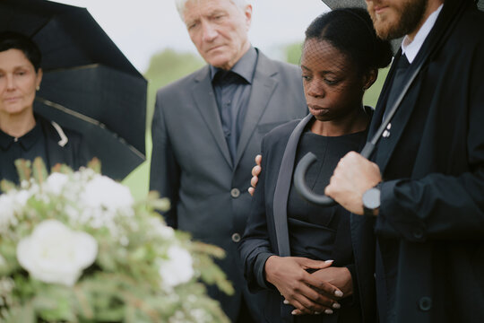 Black young adult woman standing with eyes closed being comforted by Caucasian middle aged man and woman during funeral ceremony outdoors near floral arrangement