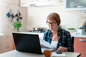 Senior woman working from home with laptop, finances, paying utility bills on kitchen. Personal...