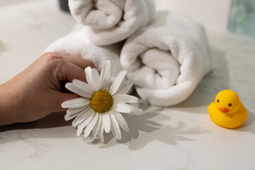 A hand gently holds a white flower next to neatly rolled towels and a yellow rubber duck. The atmosphere suggests a calming environment perfect for self-care and relaxation.