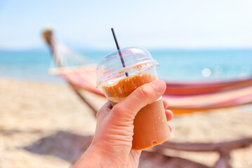 Hand holding iced coffee in a takeaway cup on a sunny beach, colorful hammock blurred in the background