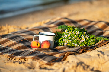 Still life on checkered plaid with metal cup, bouquet of daisies and peaches on sand in cozy summer beach setting
