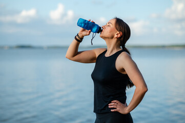 Beautiful woman in tight sportswear drinking water from sports bottle during lakeside beach run on sunny day