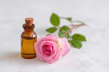 Amber glass bottle of rose essential oil sits beside a single, light pink rose with stem and leaves on a white textured background
