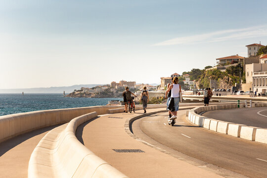 Des promeneurs sur la corniche Kennedy &agrave; Marseille, &agrave; trottinette, &agrave; v&eacute;lo et &agrave; pieds sur la piste cyclable en bord de mer avec la ville en arri&egrave;re-plan, Marseille, Sud de la France