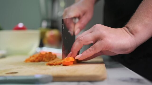 Close up of a person slicing raw carrot with a cleaver on a wooden chopping board. Home cooking scene with fresh vegetables on a kitchen counter. Preparation and healthy eating.