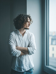 A person in a white shirt with curly hair stands by a window, looking outside thoughtfully.