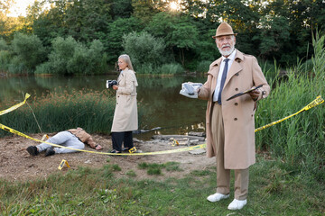Forensic expertise. Detectives working at crime scene with dead man's body near lake outdoors