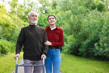 Obraz premium Smiling caregiver supporting elderly man who using walking frame outdoors. Space for text