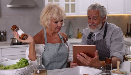 Elderly couple with tablet cooking together at table in kitchen