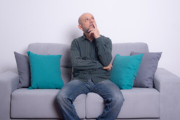 Bearded bald man wearing a long-sleeved shirt and jeans sitting on a gray sofa posing and looking...