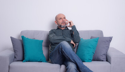 Bearded bald man wearing a long-sleeved shirt and jeans sitting on a gray sofa posing and looking...