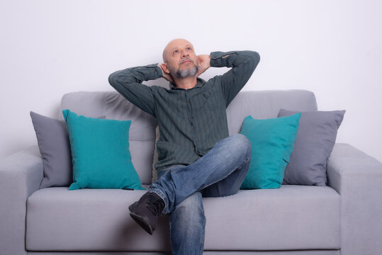 Bearded bald man wearing a long-sleeved shirt and jeans sitting on a gray sofa posing and looking up. - Powered by Adobe