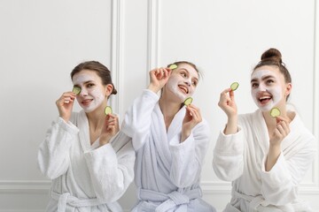 Happy women with facial masks and cucumber slices in bathrobes after spa procedures near white wall