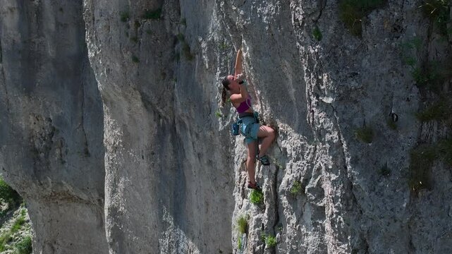 AERIAL: Young woman is focused on her climb with body positioned against sheer rock face. Wearing harness and secured by rope, she is lead climbing a challenging route in textured limestone rock wall.