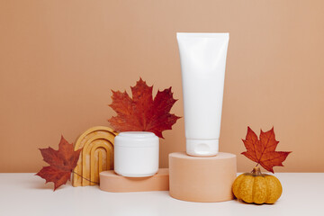 White mockup containers of body or facial cream on podium with autumn leaves and pumpkin. Skincare products for dry skin