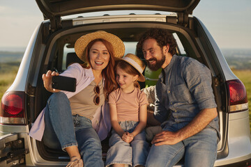 A family shares a joyful moment during an outdoor picnic, taking a selfie in the back of their car while surrounded by nature, creating lasting memories together on a sunny day.