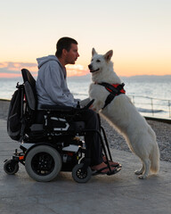Man with disability sits in a wheelchair at a waterfront. His white dog stands beside him, both...