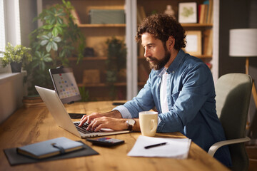 A man types on his laptop in a cozy home office. The workspace features wooden furniture, indoor...