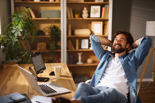 A man sits comfortably in his home office, smiling and resting his hands behind his head. Natural light fills the cozy space, highlighting a desk with a laptop and a cup.